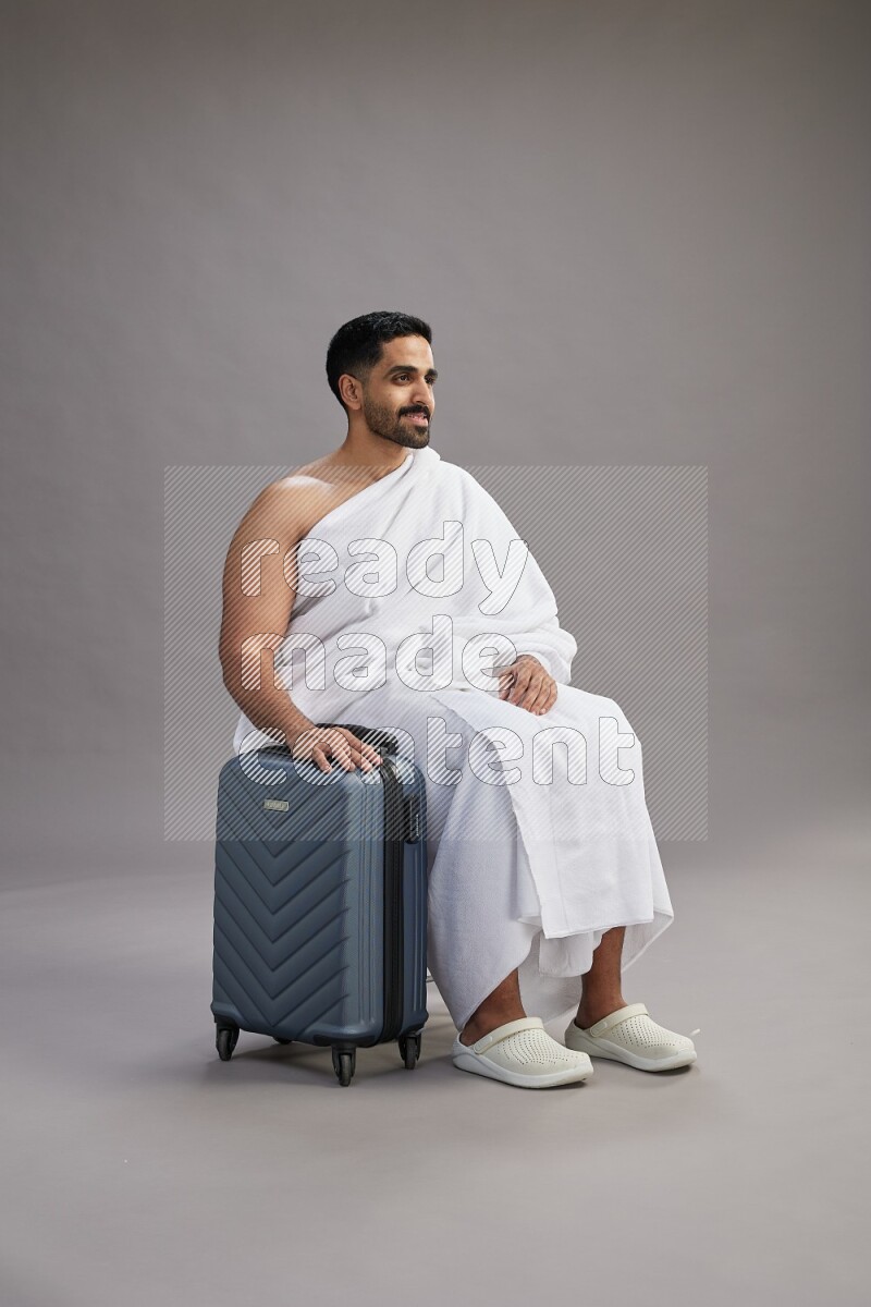 A man wearing Ehram sitting on chair holding traveling bag on gray background