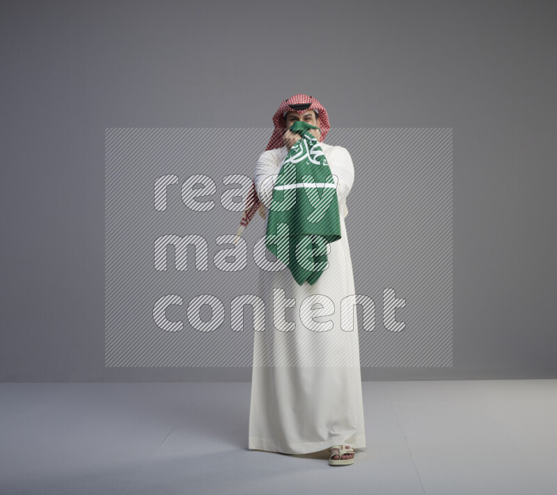 A Saudi man standing wearing thob and red shomag kissing big Saudi flag on gray background
