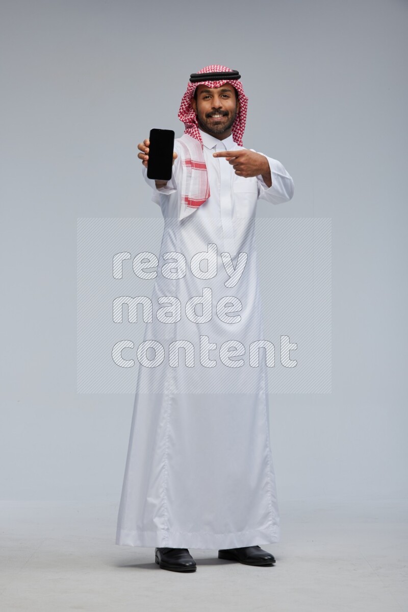 Saudi man Wearing Thob and shomag standing showing phone to camera on Gray background