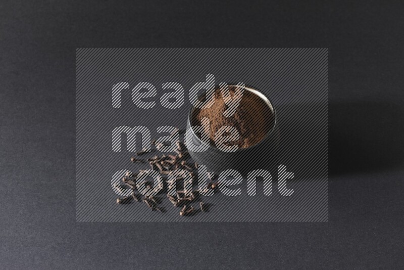 A black pottery bowl full of cloves powder and cloves whole grains on a black flooring