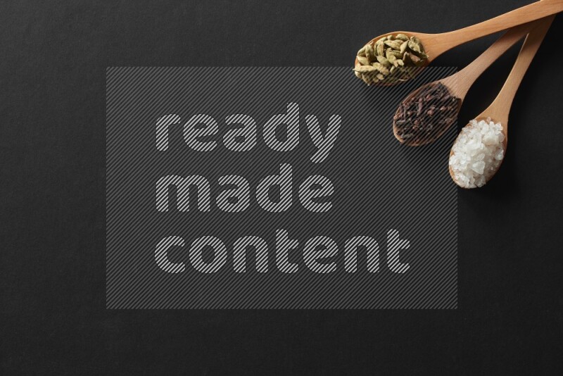 الفلفل الأبيض والقرنفل والهيل والملح والفلفل الأسود وريحان مجفف في معالق خشبية علي خلفية سوداء