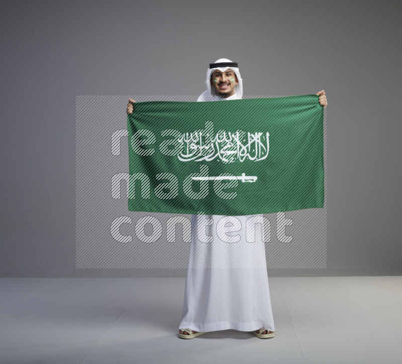 A Saudi man standing wearing thob and white shomag with face painting holding big Saudi flag on gray background