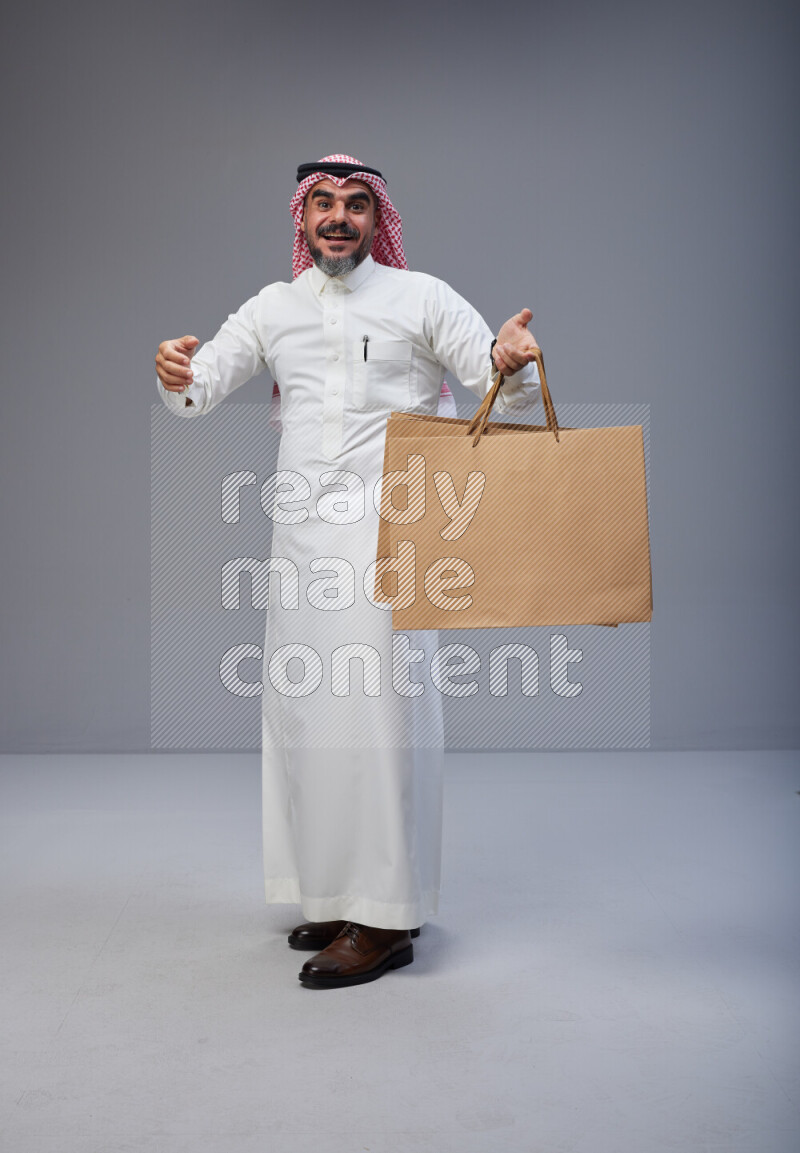 Saudi man Wearing Thob and red Shomag standing holding shopping bag on Gray background
