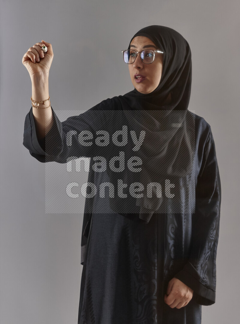 Woman in black abaya, hijab and glasses holding a marker pen to write on a transparent board with different reactions and expressions on grey background