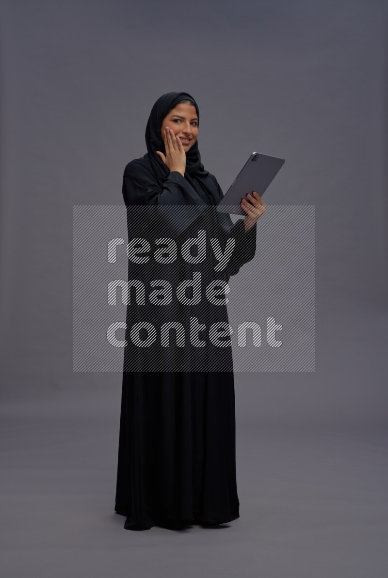Saudi woman wearing Abaya standing working on tablet on gray background