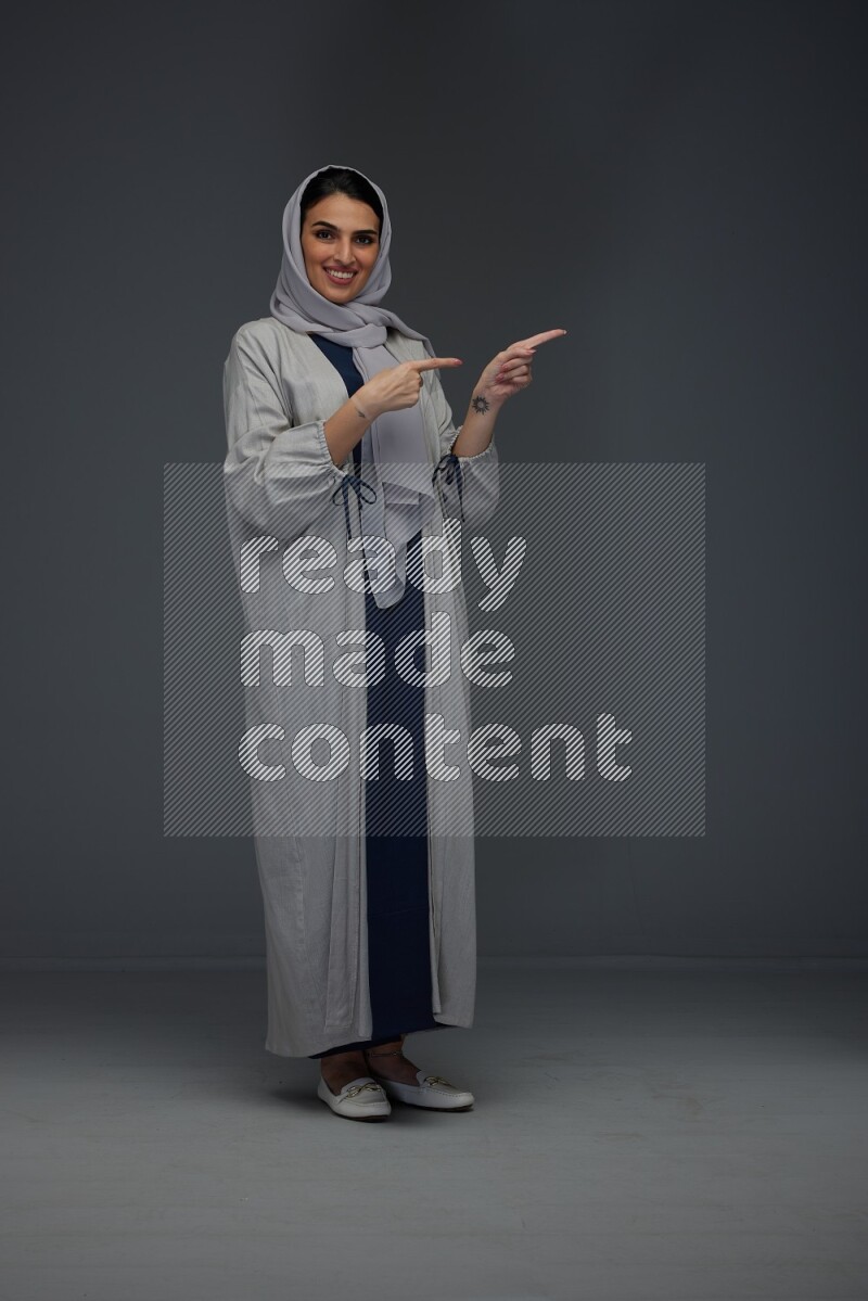 A Saudi woman wearing a light gray Abaya and head scarf standing and pointing in different directions eye level on a grey background