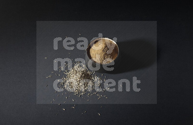 A black pottery bowl full of cumin powder and spreaded cumin seeds on a black flooring