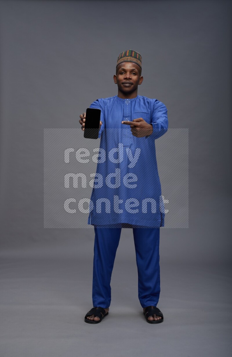 Man wearing Nigerian outfit standing showing phone to camera on gray background