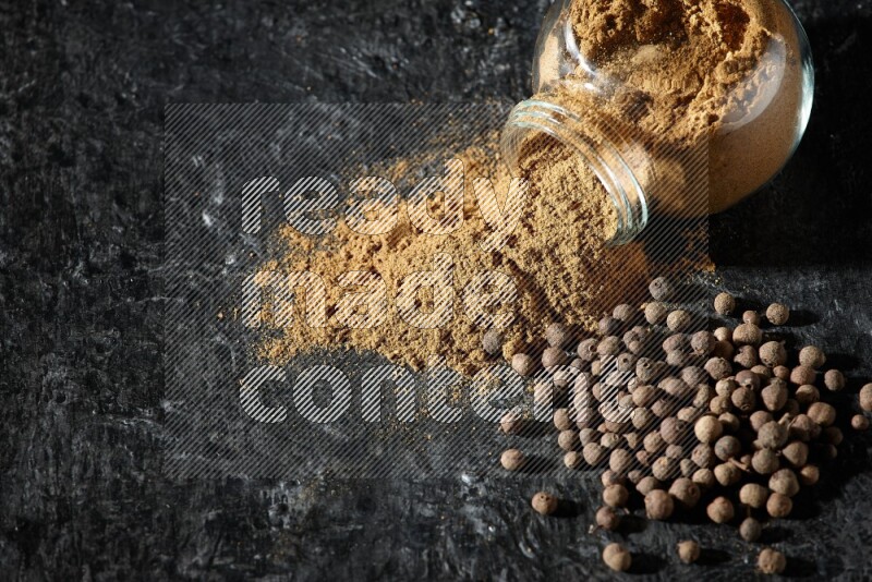 A flipped glass spice jar full of allspice powder and powder spilled out of it with whole balls on a textured black flooring
