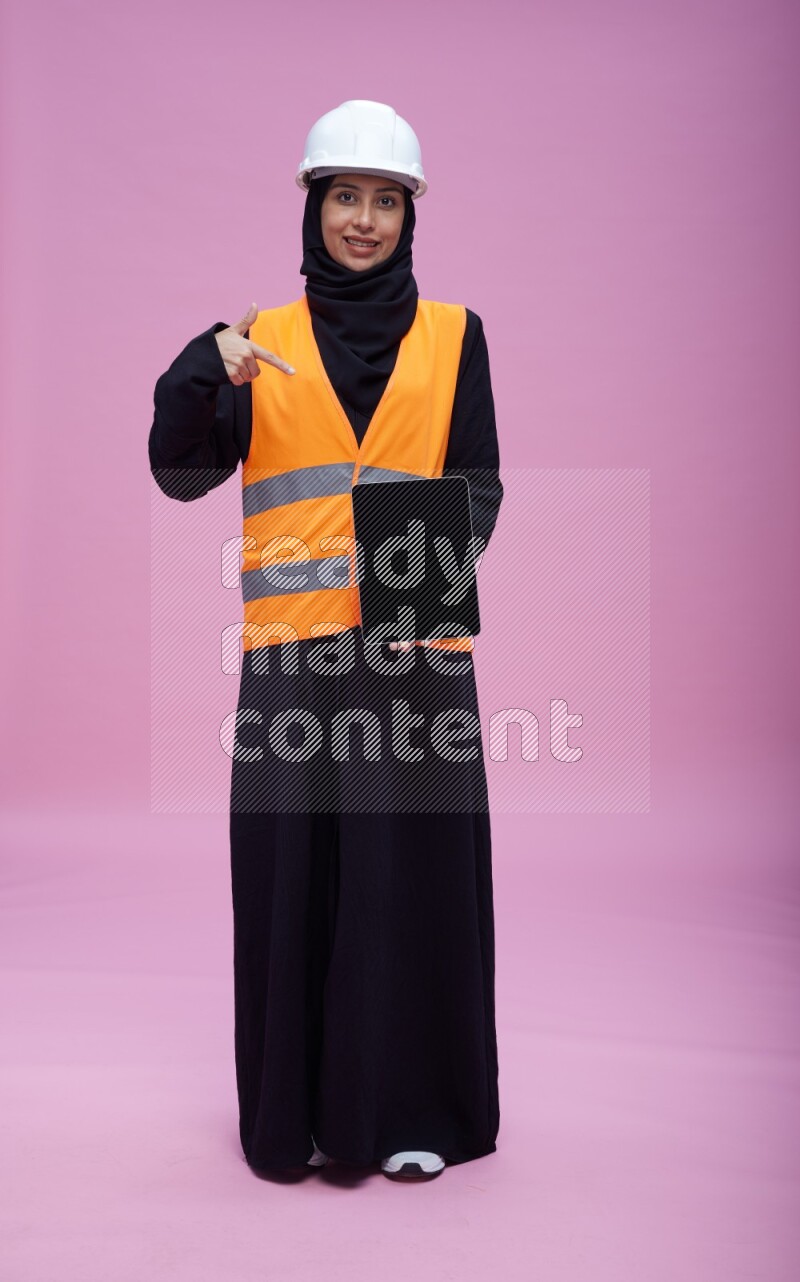 Saudi woman wearing Abaya with engineer vest and helmet standing showing tablet to camera on pink background