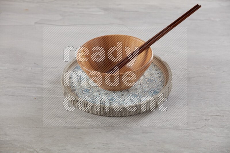 brown wood round bowl on top of multi color round ceramic plate and chopsticks, on grey textured countertop