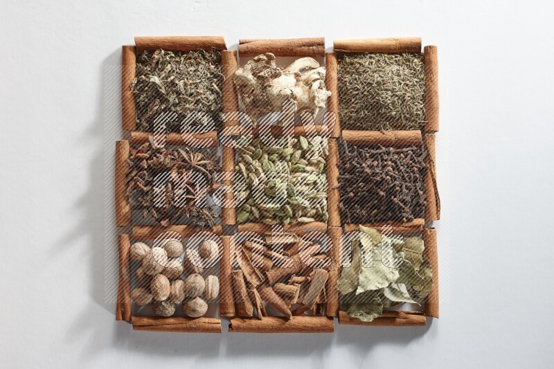 9 squares of cinnamon sticks full of cardamom in the middle surrounded by nutmeg, cinnamon, bay laurel leaves, cloves, cumin, dried ginger, dried basil and star anise on white flooring