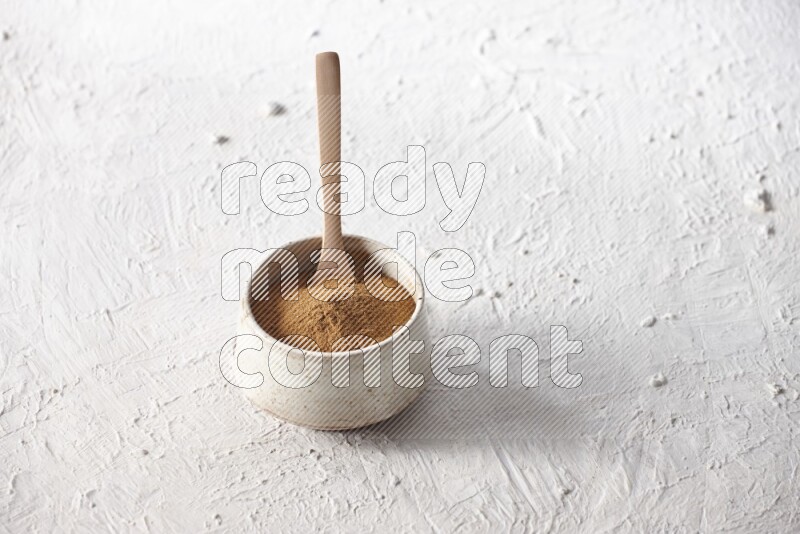 Ceramic beige bowl full of cinnamon powder with a wooden spoon on a textured white background