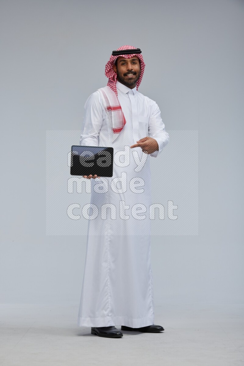 Saudi man Wearing Thob and shomag standing showing tablet to camera on Gray background