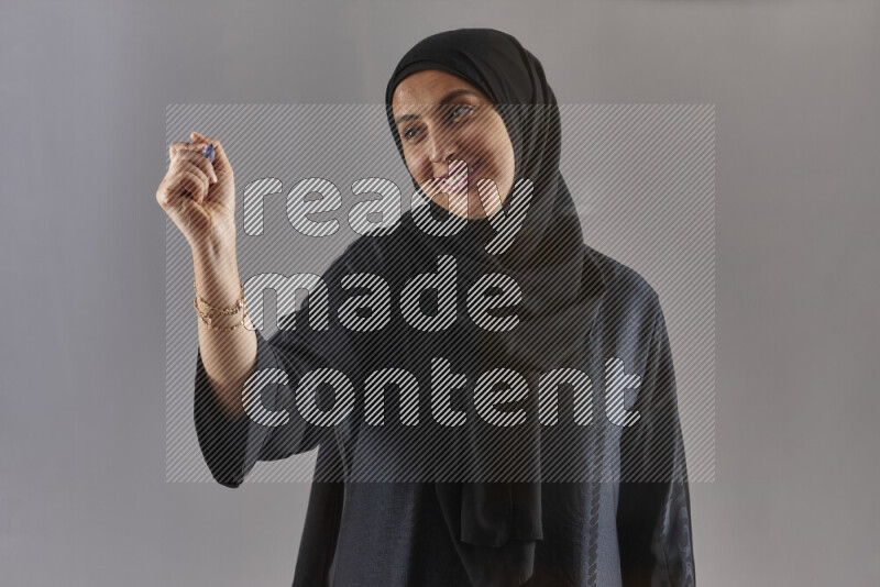 Woman in black abaya and hijab holding a marker pen to write on a transparent board with different reactions and expressions on grey background