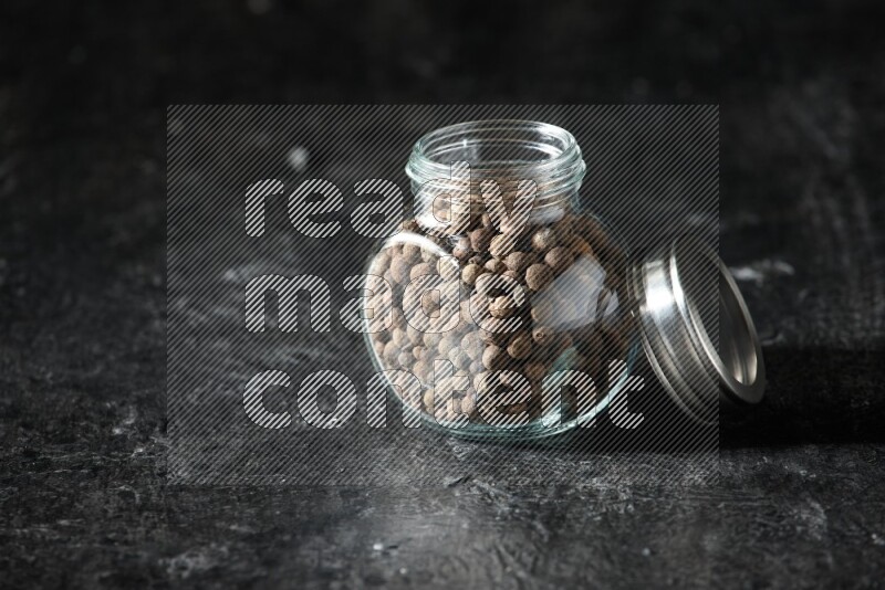 A glass spice jar full of allspice whole balls on a textured black flooring