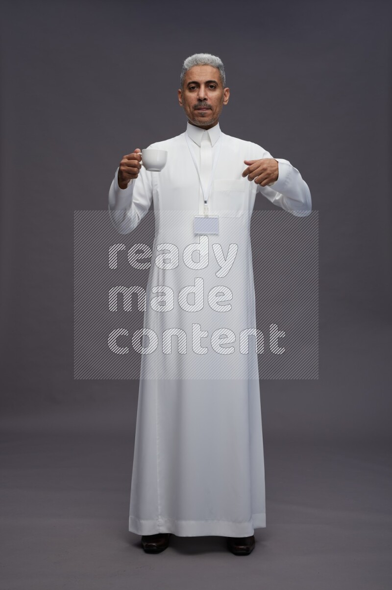 Saudi man wearing thob with neck strap employee badge standing holding mug on gray background