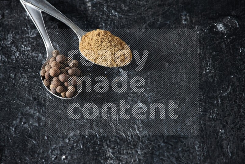 2 metal spoons full of allspice powder and whole balls on a textured black flooring