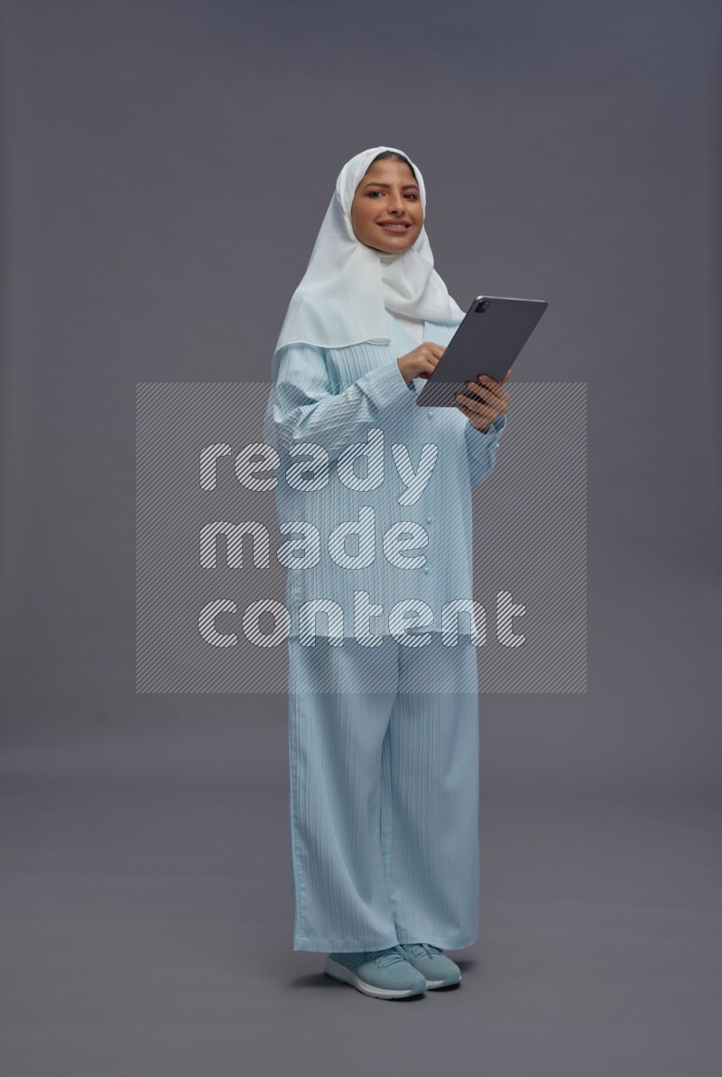Saudi woman wearing hijab clothes standing working on tablet on gray background