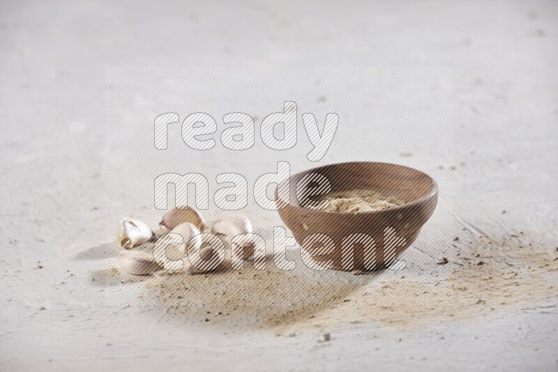 A wooden bowl full of garlic powder and beside it garlic cloves on a textured white flooring in different angles
