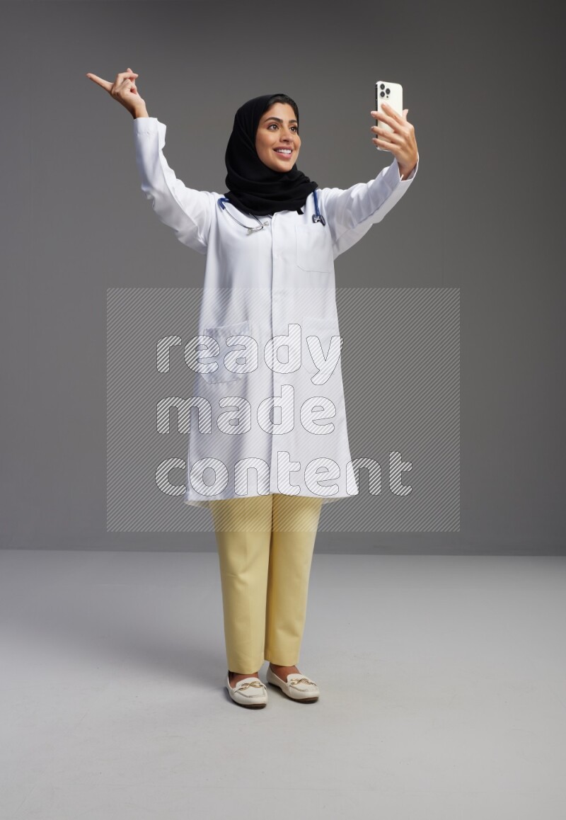 Saudi woman wearing lab coat with stethoscope standing taking selfie on Gray background