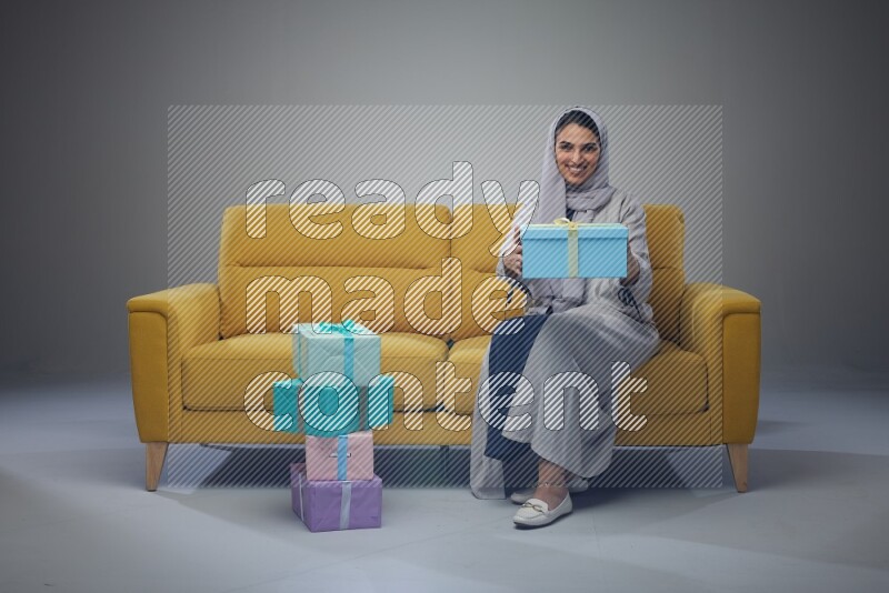 A Saudi woman wearing a light gray Abaya and head scarf sitting on a yellow sofa holding a gift box beside multi color gift boxes eye level on a grey background