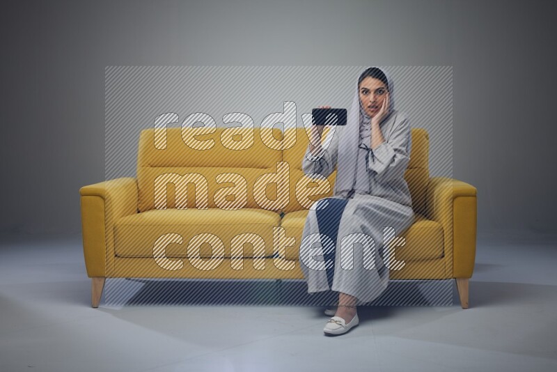 A Saudi woman wearing a light gray Abaya and head scarf setting on a yellow sofa and holding her phone while showing it's screen eye level on a grey background