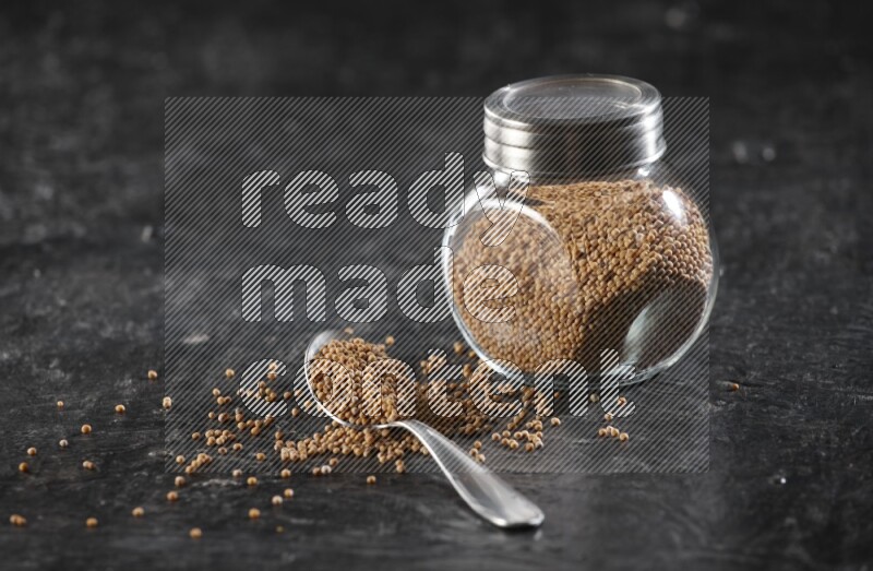 A glass spice jar and a metal spoon full of mustard seeds on a textured black flooring