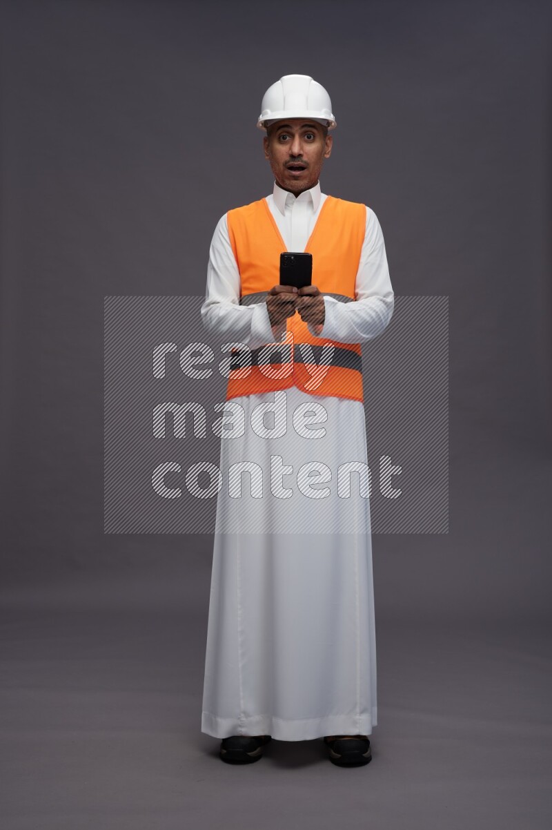 Saudi man wearing thob with engineer vest standing texting on phone on gray background