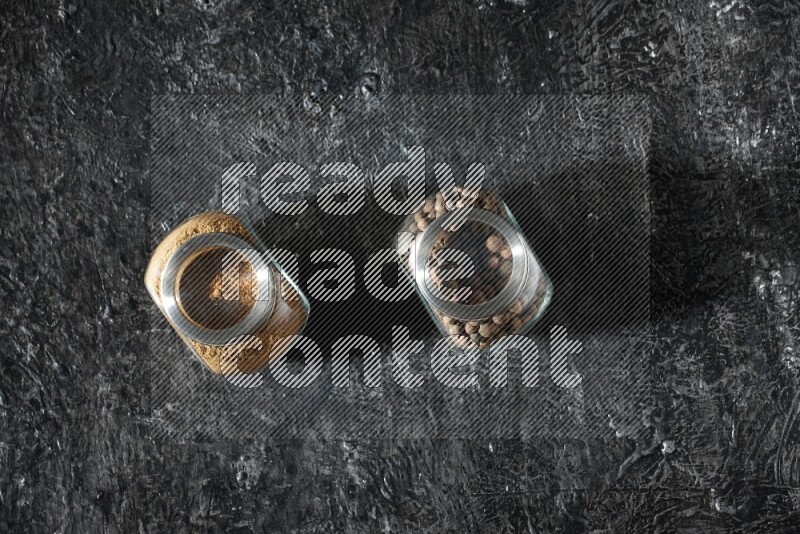 وعاءان زجاجيان للتوابل ممتلئان ببودرة بهار الفلفل الحلو وحبوبه على خلفية سوداء