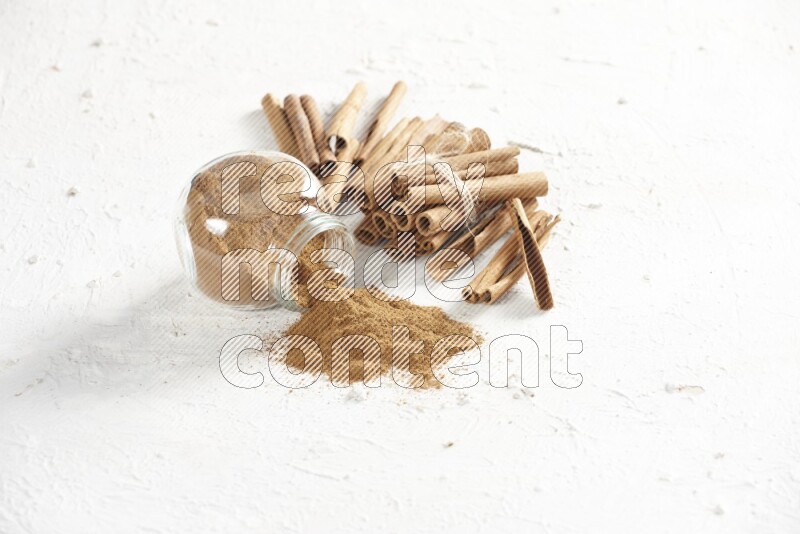 Flipped herbs glass jar full of cinnamon powder and cinnamon sticks in the back on a textured white background