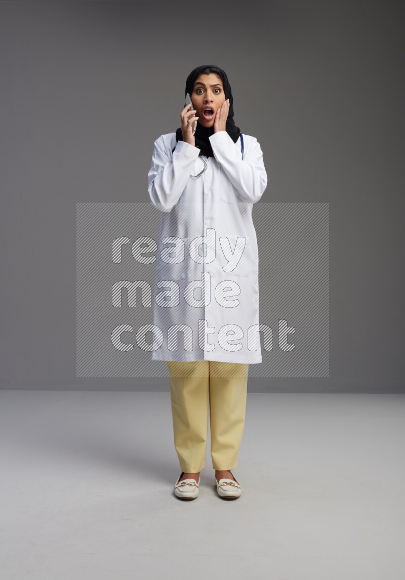 Saudi woman wearing lab coat with stethoscope standing talking on phone on Gray background