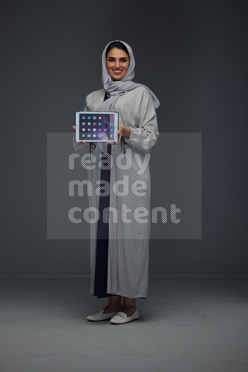 A Saudi woman wearing a light gray Abaya and head scarf standing and showing the phone's screen eye level on a grey background