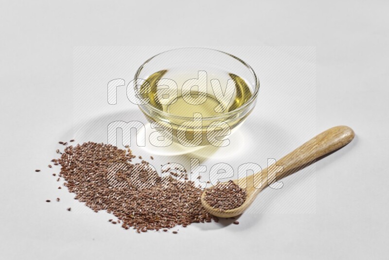 A glass bowl full of flaxseeds oil and wooden spoon full of flaxseeds with spread seeds on a white flooring