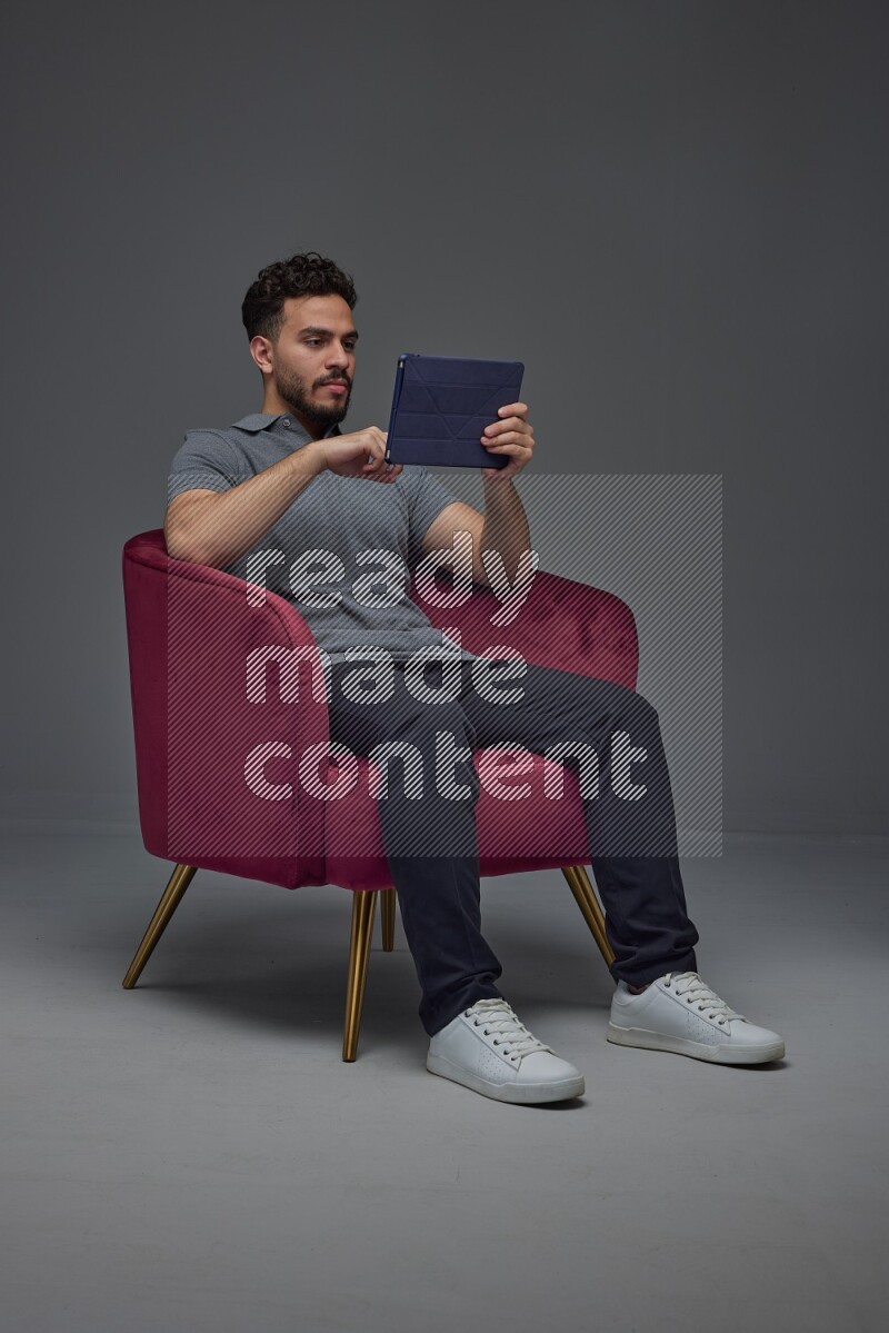 A man wearing casual and using his tablet while sitting on a burgundy chair eye level on a gray background