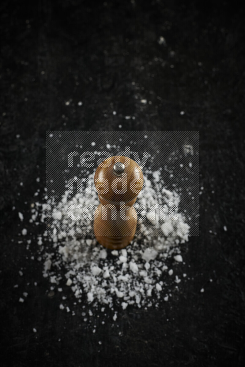 A wooden grinder standing upright and surrounded by coarse white sea salt on black background