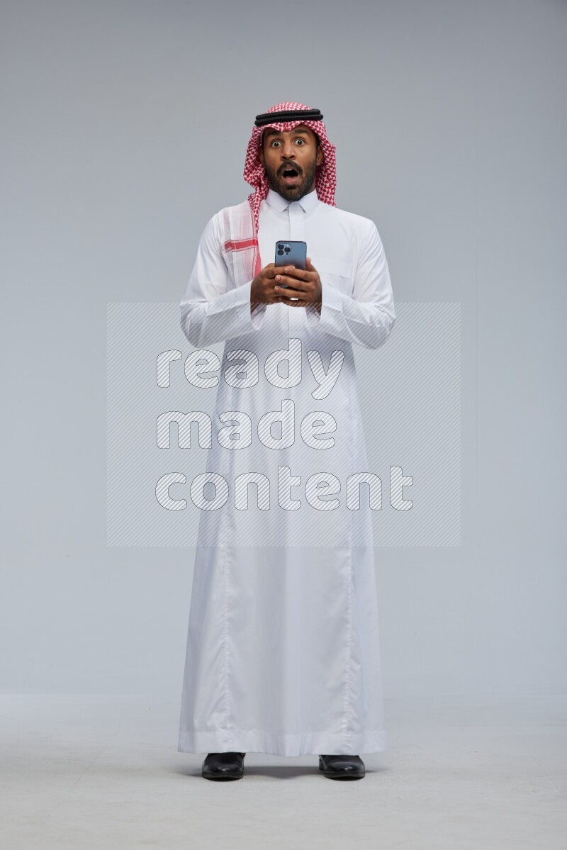 Saudi man Wearing Thob and shomag standing texting on phone on Gray background
