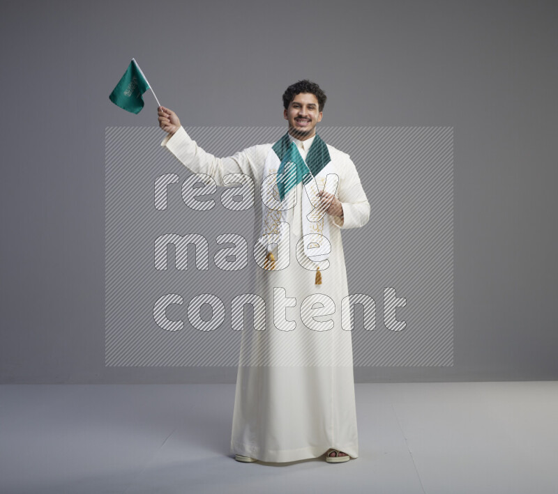 A Saudi man standing wearing thob and saudi flag scarf and holding small saudi flag on gray background