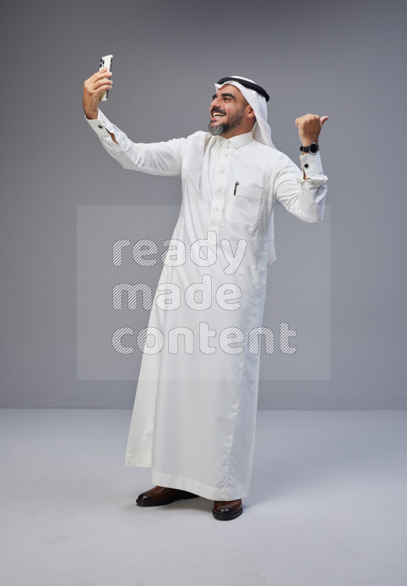 Saudi man Wearing Thob and white Shomag standing taking selfie on Gray background