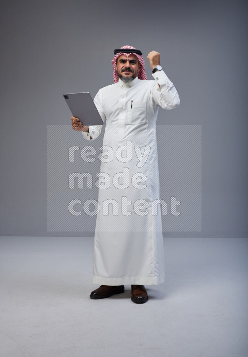 Saudi man Wearing Thob and red Shomag standing working on tablet on Gray background