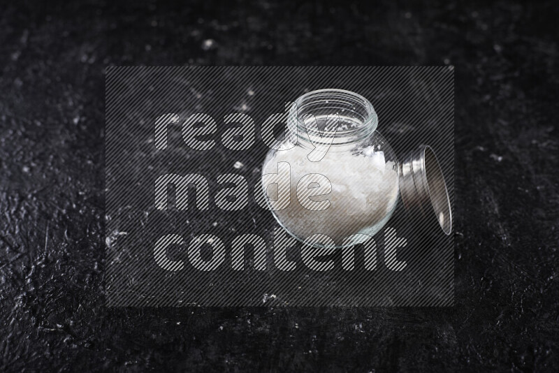A glass jar full of coarse sea salt crystals on black background