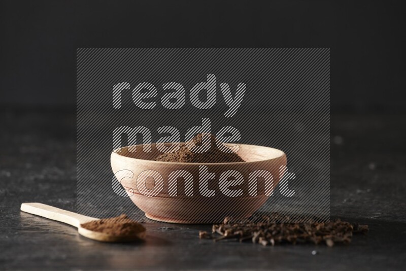 A wooden bowl and a wooden spoon full of cloves powder with spreaded cloves on a textured black flooring