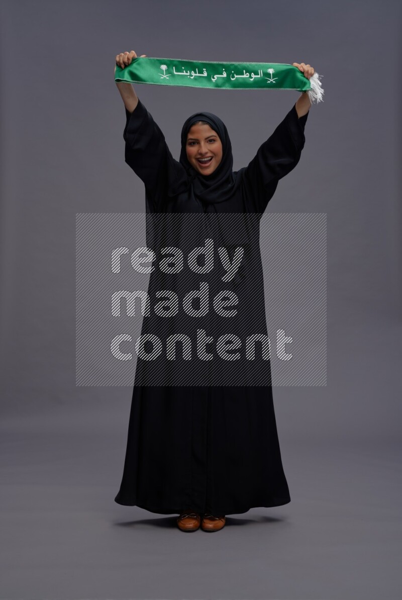 Saudi woman wearing Abaya standing holding Saudi flag on gray background