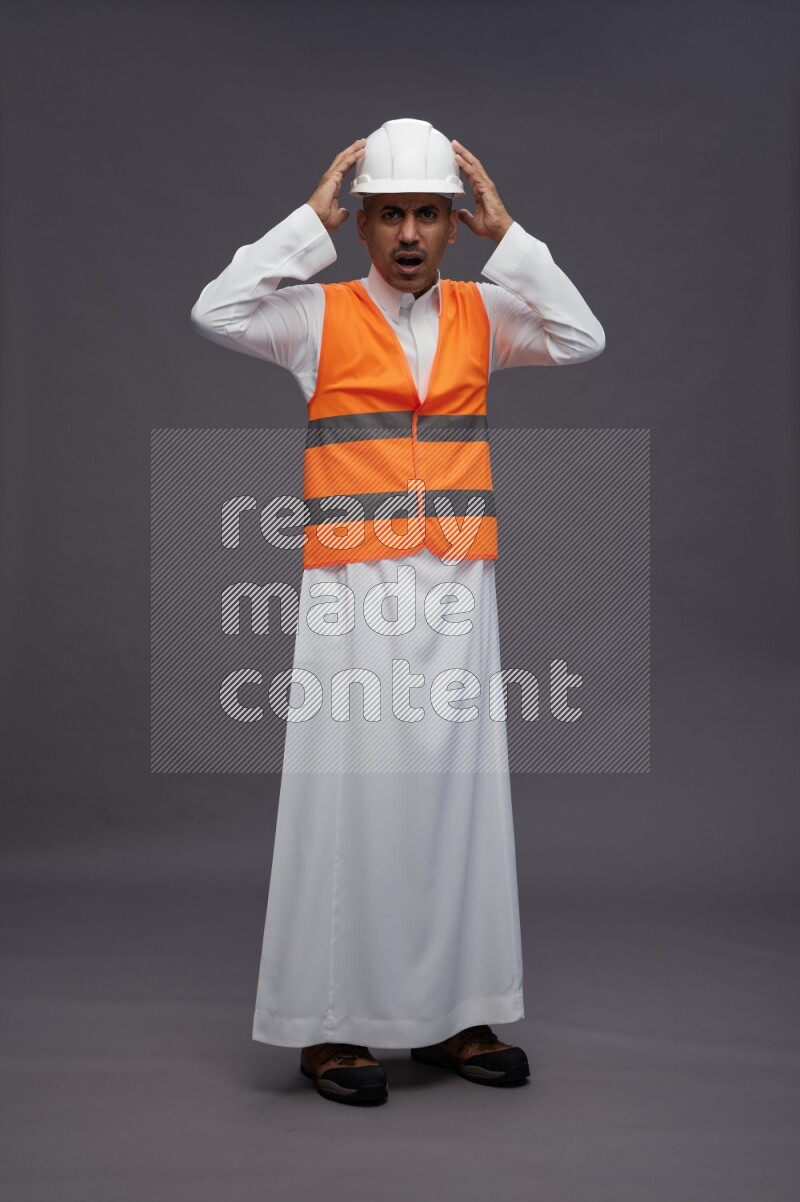 Saudi man wearing thob with engineer vest standing hands on head on gray background