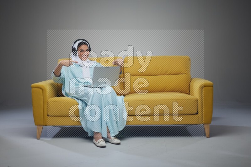A Saudi woman sitting on a yellow Sofa With her Laptop Pointing wearing Blue Abaya