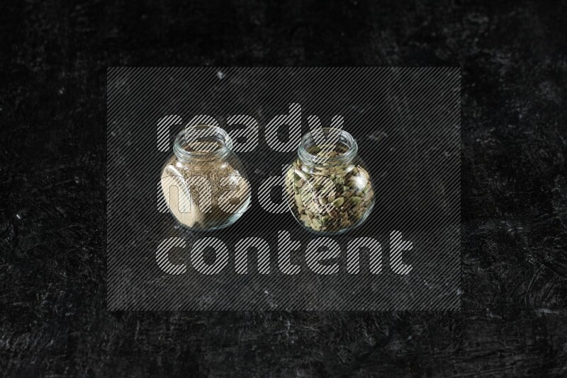 2 glass spice jars full of cardamom powder and cardamom seeds on textured black flooring