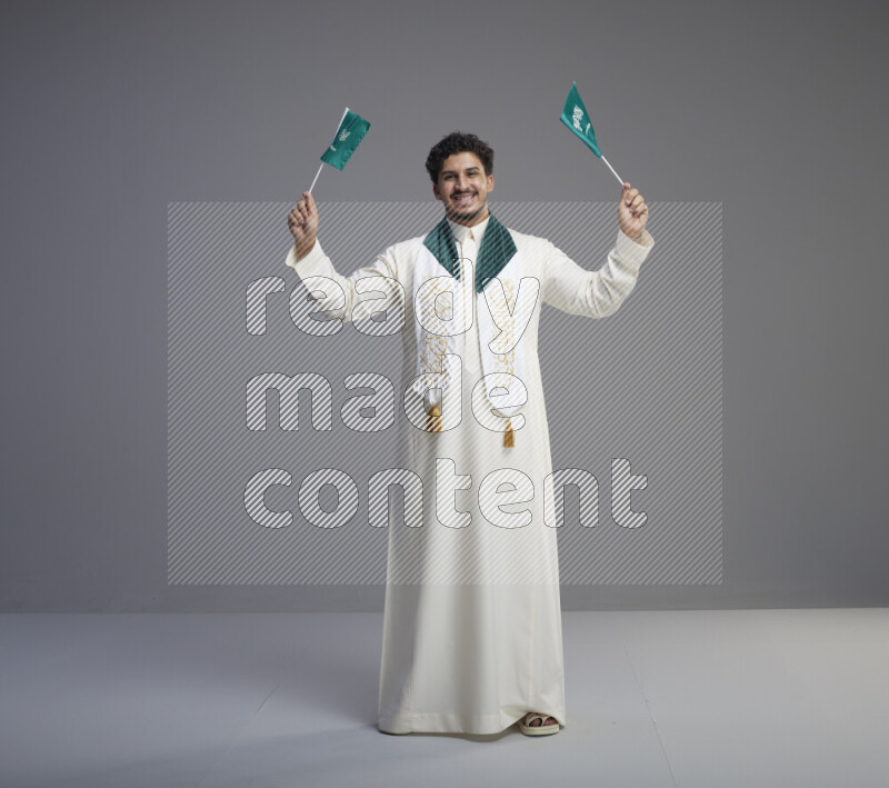 A Saudi man standing wearing thob and saudi flag scarf and holding small saudi flag on gray background