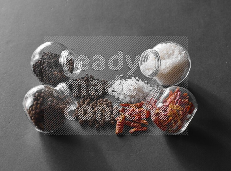 4 glass spice jars full of salt, chili peppers, black peppers and allspice on black flooring