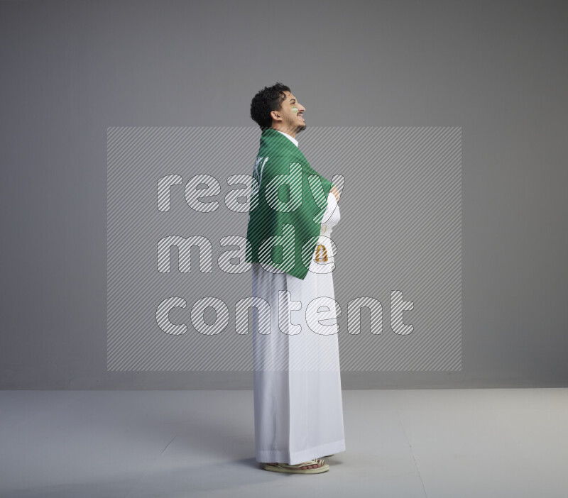 A Saudi man standing wearing thob and saudi flag scarf with face painting wrapping big Saudi flag on gray background