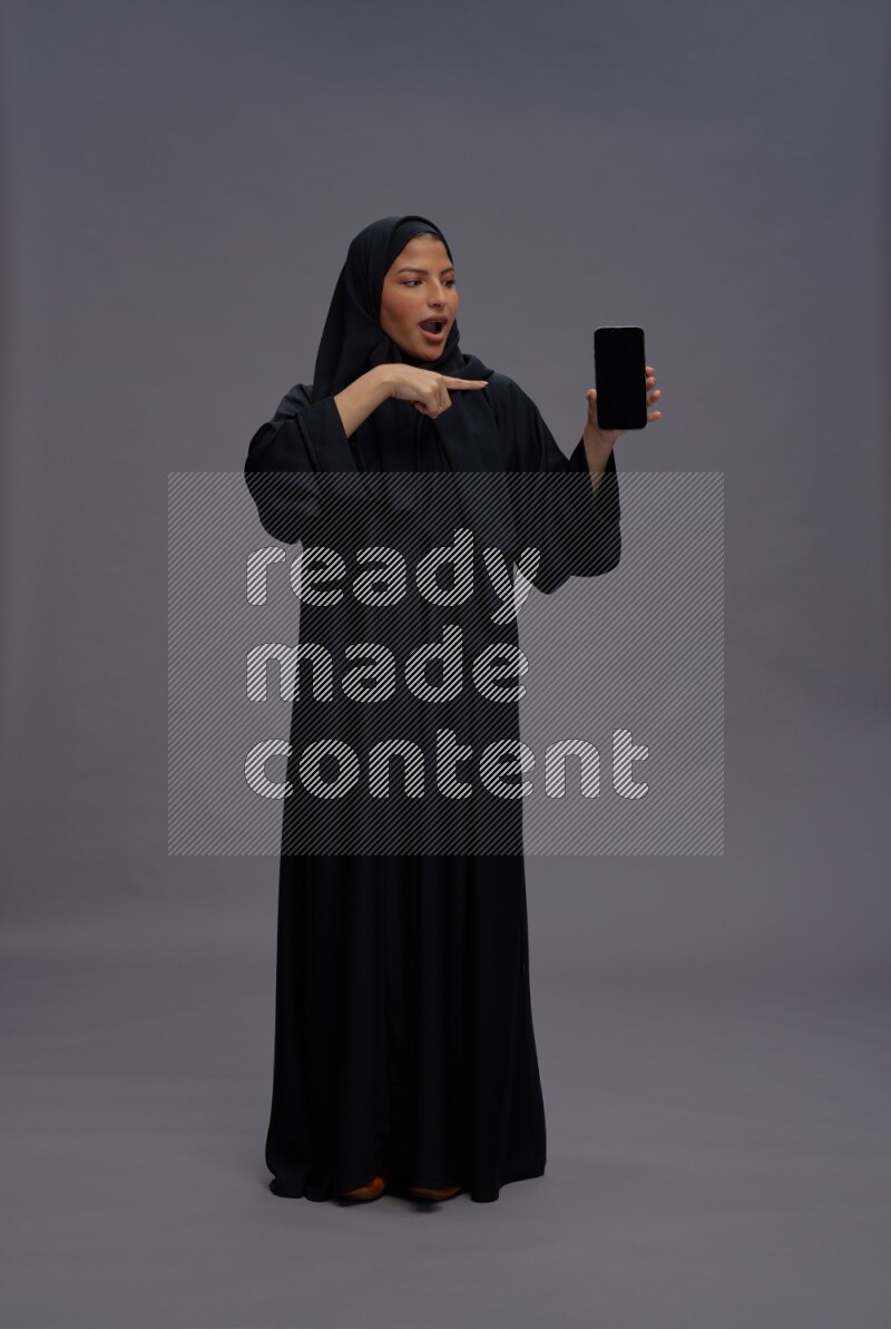 Saudi woman wearing Abaya standing showing phone to camera on gray background